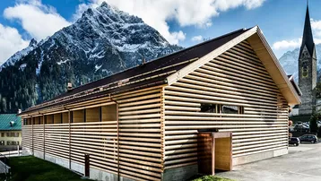 Modernes Gebäude mit Holzfassade vor einer beeindruckenden Bergkulisse und einem Kirchturm im Hintergrund.