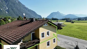 Gelbes Gebäude mit braunem Dach in alpiner Landschaft mit Bergblick und grünen Wiesen.
