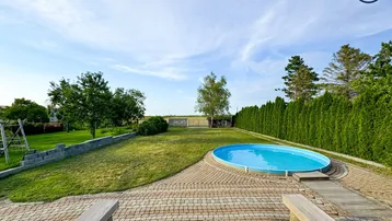 Großer Gartenbereich mit Rasenfläche, ovalem Pool und gepflasterter Terrasse mit Sitzbänken.