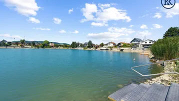 Seeufer mit Häusern im Hintergrund und Zugang zum Wasser unter blauem Himmel.