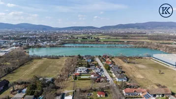 Luftaufnahme einer Wohnsiedlung am Seeufer mit umliegender Landschaft und Bergen im Hintergrund.