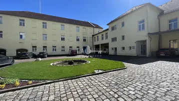 Außenansicht eines Gebäudekomplexes mit gepflastertem Innenhof, Grünfläche und einem kleinen Brunnen.