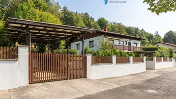 Hausansicht von der Straße mit Carport und Holzzaun, umgeben von viel Grün.