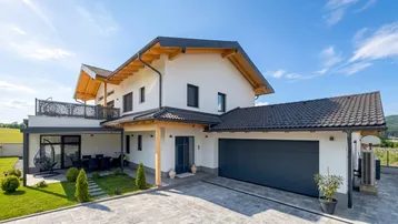 Modernes Einfamilienhaus mit weißer Fassade, dunklem Dach und integrierter Garage unter blauem Himmel.