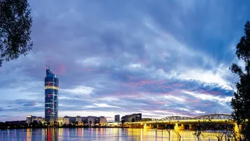 Abenddämmerung über der Donau mit beleuchteter Skyline und Brücke, die sich im Wasser spiegelt.