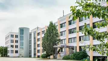 Modernes Bürogebäude mit Glasfassade und gepflegter Außenanlage unter bewölktem Himmel.