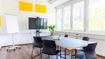 Moderner Konferenzraum mit großem Tisch, Bildschirm und Flipchart, ideal für Besprechungen und Präsentationen.