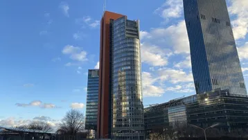 Moderne Hochhäuser mit Glasfassaden und einem roten Turm unter blauem Himmel, umgeben von städtischer Infrastruktur.