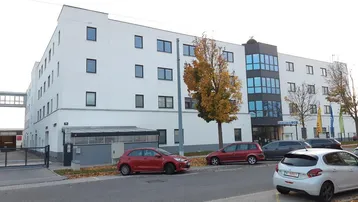 Modernes Bürogebäude mit weißer Fassade, blauen Glasfenstern und parkenden Autos auf der Straße.