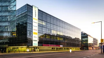 Modernes Bürogebäude mit Glasfassade und beleuchteten Werbetafeln an einer belebten Straße bei Sonnenuntergang.