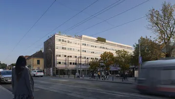 Gebäudeansicht von der Straße mit vorbeifahrender Straßenbahn und städtischer Umgebung an einem sonnigen Tag.