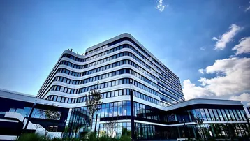 Modernes Bürogebäude mit geschwungener Architektur und vielen Glasflächen unter blauem Himmel.
