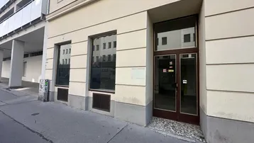 Die Fassade des Gebäudes zeigt eine Ladenfront im Erdgeschoss mit großen Schaufenstern und einem Eingang.