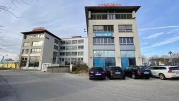 Modernes Bürogebäude mit mehreren Etagen und großen Fenstern, umgeben von einem Parkplatz unter klarem Himmel.