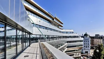 Moderne Architektur mit geschwungener Glasfassade und einer langen Terrasse mit Stadtblick unter blauem Himmel.