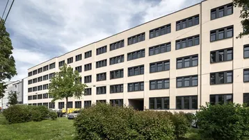 Modernes Bürogebäude mit vielen Fenstern und gepflegter Grünanlage davor unter bewölktem Himmel.