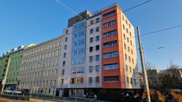 Modernes Gebäude mit mehreren Etagen, großen Fenstern und farbigen Akzenten an der Fassade, gelegen an einer Straße mit Straßenbahnschienen.
