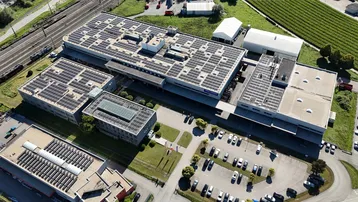 Luftaufnahme eines großen Industriekomplexes mit mehreren Gebäuden, Solaranlagen auf den Dächern und weitläufigen Parkplätzen.