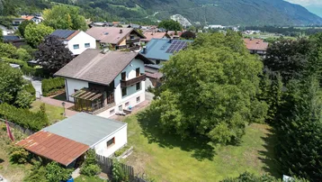 Luftaufnahme eines Einfamilienhauses mit großem Garten und umliegender Wohnsiedlung in bergiger Landschaft.