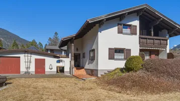 Traditionelles Einfamilienhaus im alpinen Stil mit Garage und Gartenbereich vor einer Bergkulisse.
