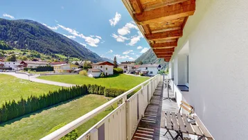 Sonniger Balkon mit Sitzgelegenheiten und weitem Blick über grüne Wiesen und Berge.