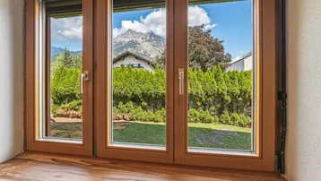 Fenster mit Blick auf den gepflegten Garten, eine Hecke und die umliegenden Berge.