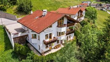 Traditionelles Alpenhaus mit rotem Ziegeldach und mehreren Balkonen, eingebettet in eine grüne Berglandschaft.
