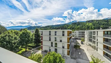 Blick auf moderne Wohngebäude mit grüner Umgebung und Bergen im Hintergrund unter blauem Himmel.