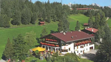 Luftaufnahme eines traditionellen Gebäudes mit großer Außenterrasse, umgeben von grünen Hügeln und Bergen.
