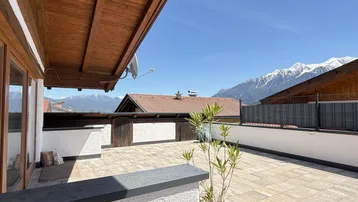 Große gepflasterte Terrasse mit Holzdachüberstand und beeindruckendem Bergblick unter blauem Himmel.