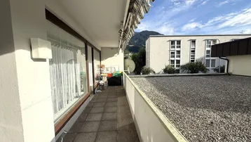 Langer Balkon mit Fliesenboden, Markise und Blick auf umliegende Gebäude und Berge.