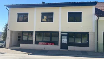 Gebäude mit hellgelber Fassade, dunklen Fensterläden und einem Eingangsbereich an einer Straße.