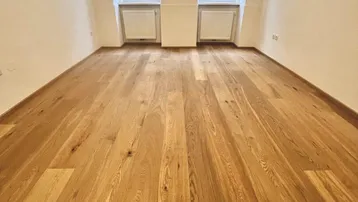 Helles Zimmer mit neuem Holzparkettboden und zwei Fenstern, die auf die Straße blicken.