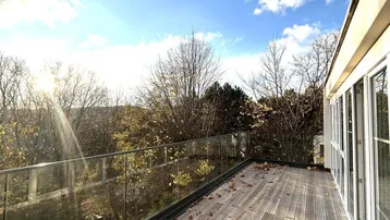 Große Holzterrasse mit Glasgeländer und Blick ins Grüne unter blauem Himmel.