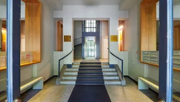 Heller Eingangsbereich mit Terrazzoboden, Treppe und modernem Glasaufzug im Hintergrund.