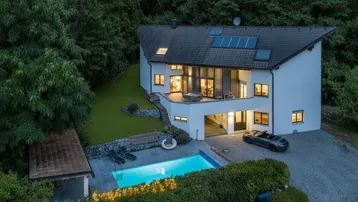 Modernes Haus mit Pool, großer Terrasse und Solaranlage, umgeben von viel Grün.