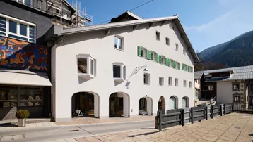 Historisches Gebäude mit weißer Fassade, grünen Fensterläden und Bergblick in einer malerischen Gasse.