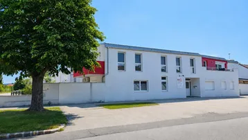 Weißes Mehrfamilienhaus mit roten Akzenten und einem großen Baum davor unter blauem Himmel.