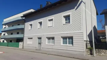 Graue Hausfassade mit Schindeln und horizontalen Paneelen an einem sonnigen Tag.
