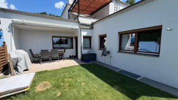 Terrasse mit Gartenbereich und Gartenmöbeln vor dem Haus unter blauem Himmel.