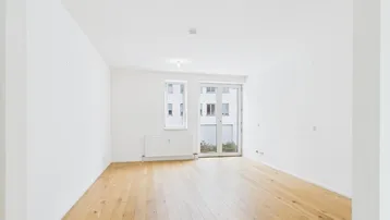 Helles, leeres Zimmer mit Holzboden und Zugang zu einem kleinen Balkon oder einer Loggia.