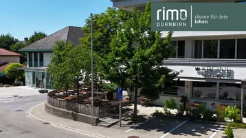 Außenansicht eines Gebäudes mit einem Restaurant und einer möblierten Terrasse unter Bäumen an einem sonnigen Tag.