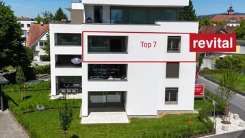 Modernes weißes Apartmentgebäude mit grüner Umgebung und Bergblick, wobei eine Einheit hervorgehoben ist.