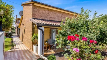 Seitlicher Blick auf das Haus mit gepflegtem Garten, blühenden Rosen und einer kleinen überdachten Terrasse.