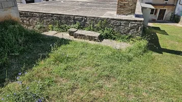 Ungenutzte Außenfläche mit einer Steinmauer und Betonplattform, umgeben von Gras und ländlicher Umgebung.