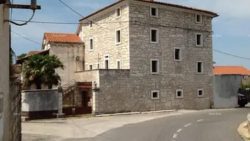Traditionelles Steinhaus mit mehreren Fenstern und rotem Ziegeldach an einer ländlichen Straße.