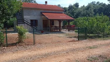 Kleines Haus mit rotem Ziegeldach, überdachter Terrasse und Außentreppe, umgeben von viel Grün in ländlicher Umgebung.