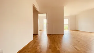Helles, leeres Wohnzimmer mit Parkettboden und Zugang zu weiteren Räumen.