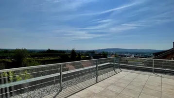 Sonnige Dachterrasse mit Fliesenboden, modernem Geländer und weitem Blick über die Landschaft und die Stadt.