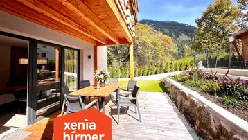 Große Holzterrasse mit Esstisch und Stühlen, umgeben von grüner Natur und Bergblick.
